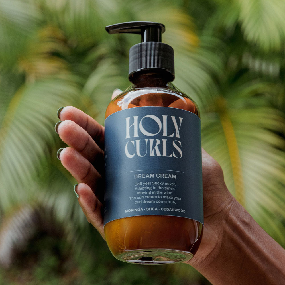 Hand holding a bottle of 'Holy Curls' Dream Cream with a blurred green leafy background