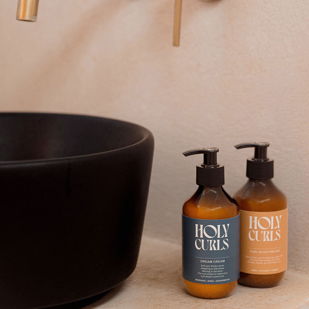 Two bottles of 'Holy Curls' products on a bathroom counter with a black bowl in the background.