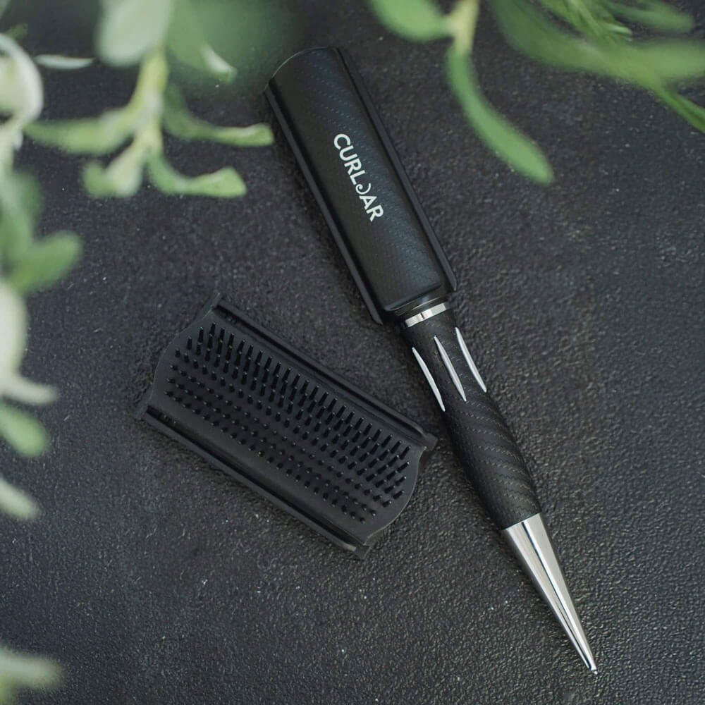 Black hair styling tool with a metallic tip on a dark textured surface with green leaves.