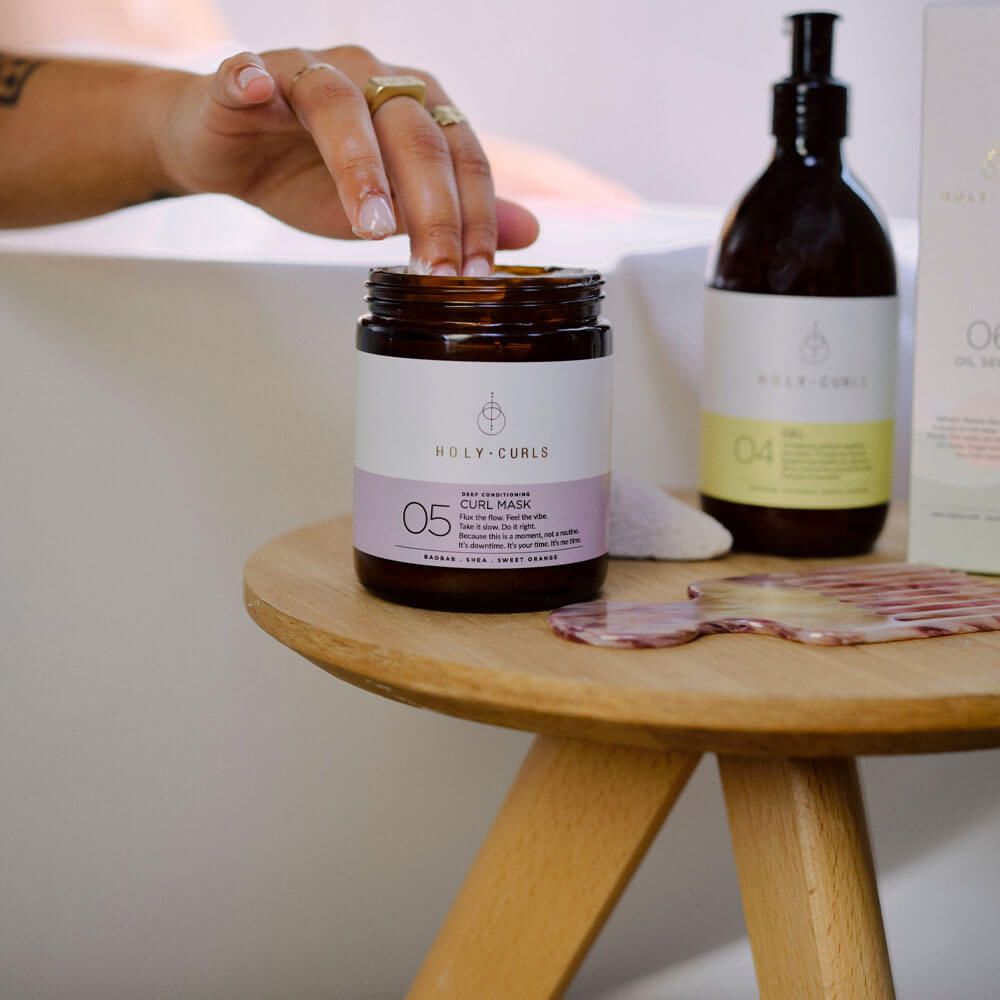 Person opening a jar of Holy Curls curl mask on a wooden table with other hair care products.