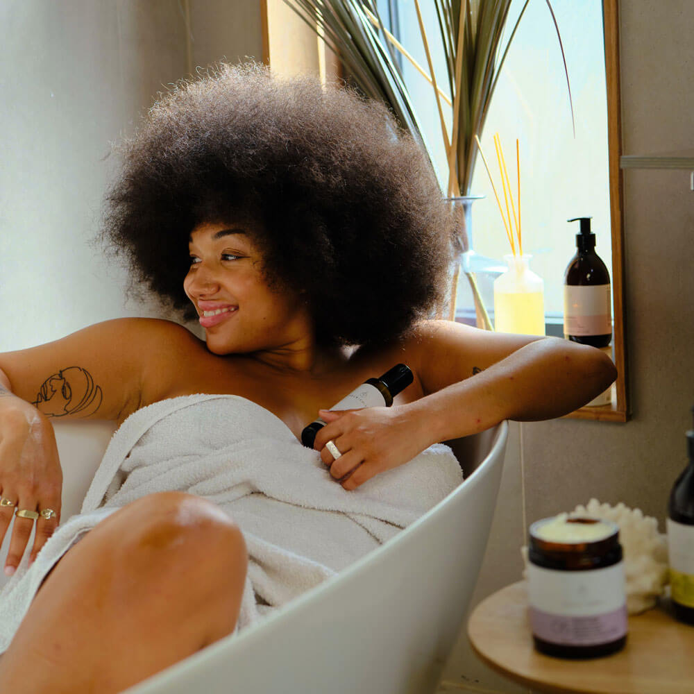 Woman with afro hair sitting in a bathtub wrapped in a towel, surrounded by bath products.