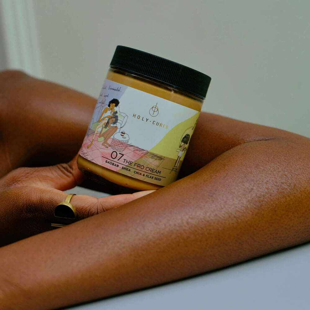 Hand holding a jar of 'Holy Curls' hair care product on a white surface