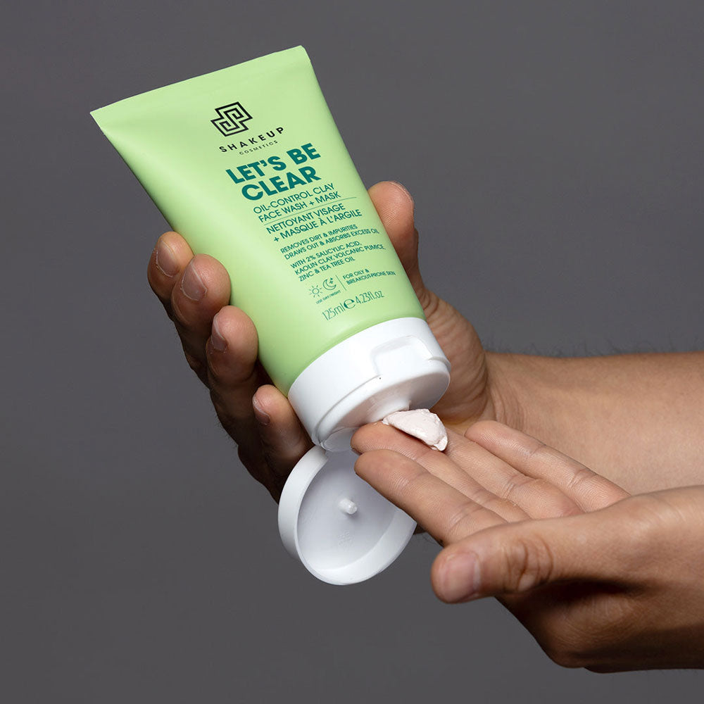 Hand holding a green skincare product labeled 'Let's Be Clear' against a gray background