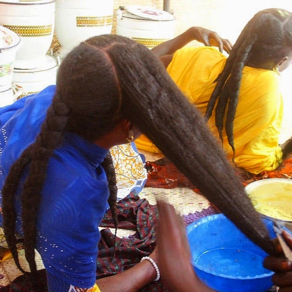 Two women with long braided hair sitting at a table with containers and a bowl using chebe.