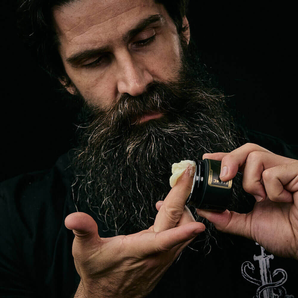 Man with a beard applying cream to a razor against a dark background