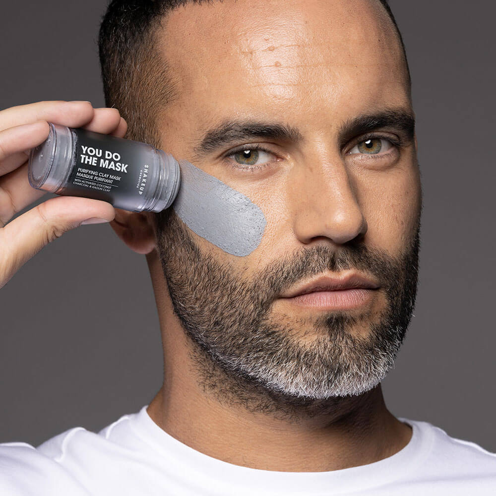 Man applying a face mask with a product labeled 'You Do The Mask' against a gray background