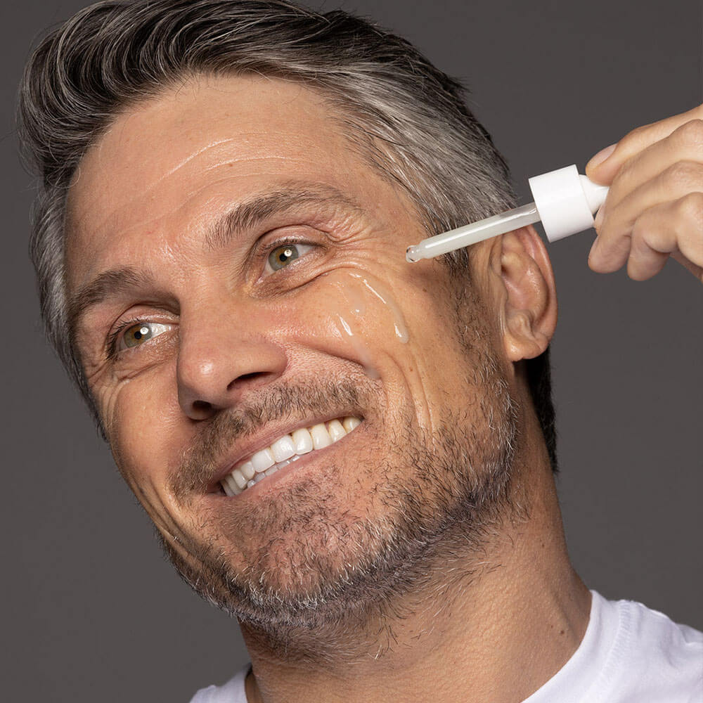 Man applying a dropper of skincare product to his face against a gray background