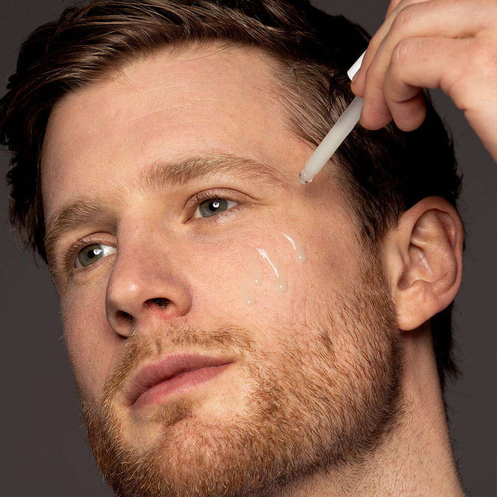Man applying skincare product to his face with a brush against a neutral background
